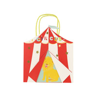 Circus themed paper party favor bag with handle in the shape of a circus tent, red and white braid stripes printed on front and back, flag at top, colorful circus sign and a lion at the center of the tent opening on front only. Enhanced with shiny gold foil stars
