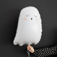 A white ghost-shaped foil balloon with a sleeping face and pink cheeks, held by a hand wearing a black and white polka dot sleeve against a dark gray background.