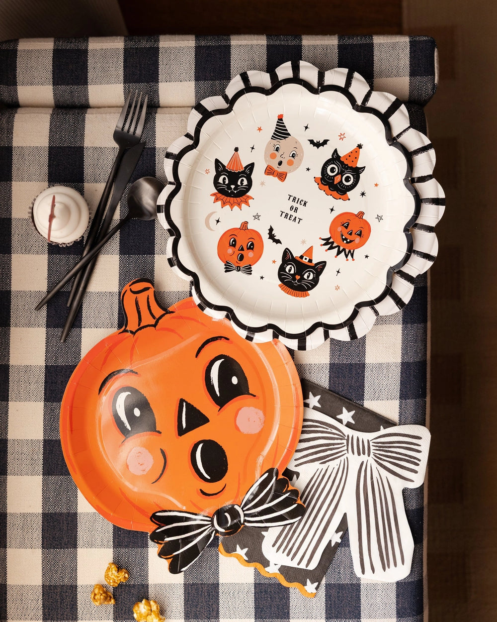 Halloween Jack-o-lantern paper party plate in the shape of a pumpkin, shown with coordinating paper plate featuring vintage characters on a black and white checkered tablecloth.