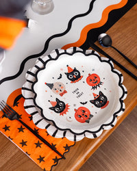 Halloween characters plate shown in a table top place setting, plate includes a printed ‘ trick or treat “ at center and is surrounded by vintage inspired characters, including moon, two cats, two jack-o-lanterns, owl, two small flying bats and stars. Cream plate with orange and black characters.