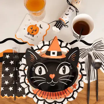 A styled Halloween table setting featuring a black cat face paper plate with an orange witch hat and ruffled collar, surrounded by black-and-white star napkins, cream beverage napkin with a orange pumpkin and trick or treat printed text, black utensils, a white mug with a brown drink, and a paper cracker party favor.