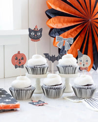 Halloween-themed cupcake toppers on white-frosted cupcakes, including a witch cat, pumpkin, bat, striped bow, ghost, and jack-o'-lantern, styled on a white table with orange and black paper fan decorations in the background.