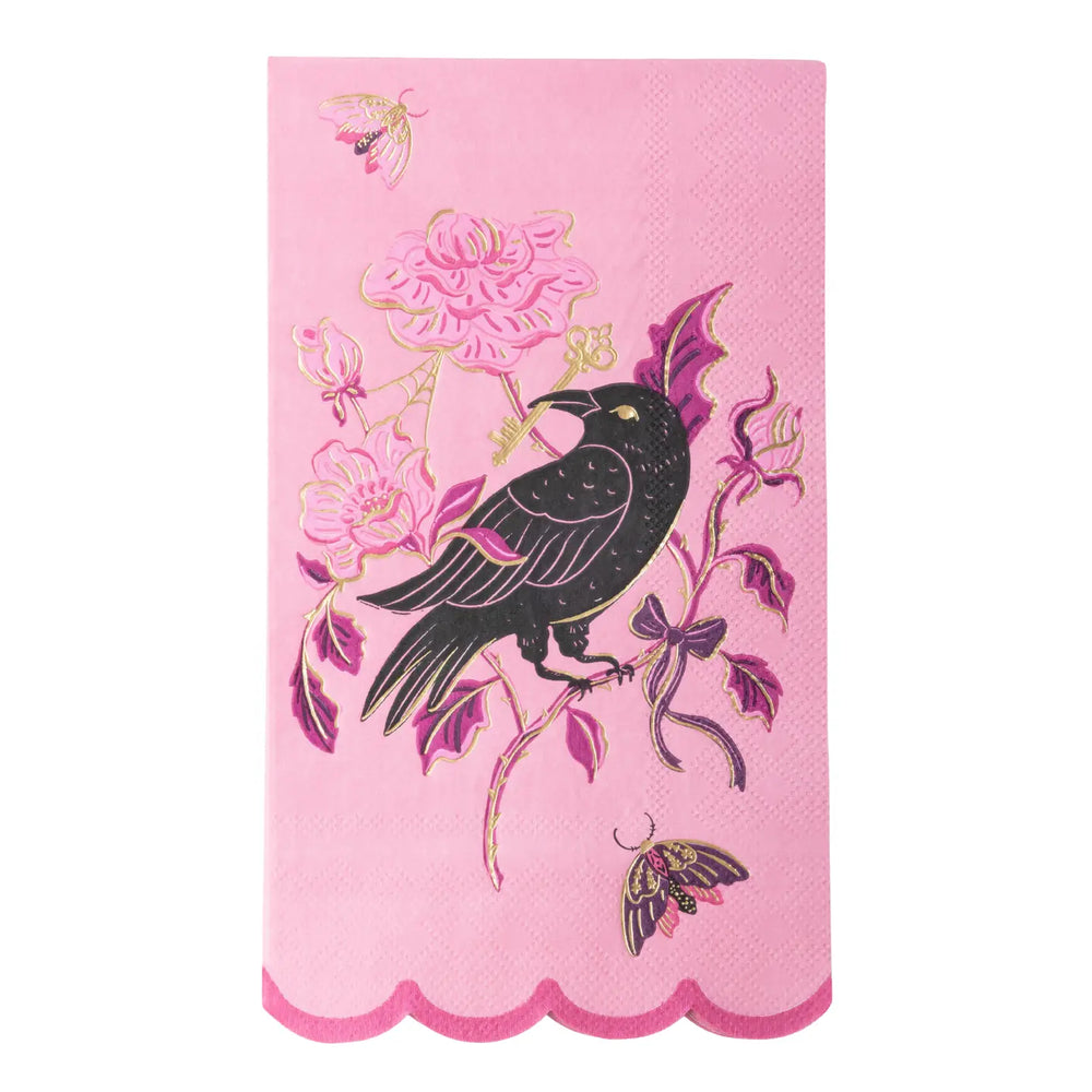 A pink decorative paper guest towel napkin featuring a black raven perched on a floral branch with pink flowers, purple leaves, and gold outlines, finished with a scalloped pink border.