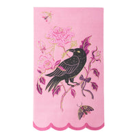 A pink decorative paper guest towel napkin featuring a black raven perched on a floral branch with pink flowers, purple leaves, and gold outlines, finished with a scalloped pink border.