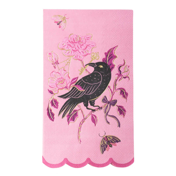 A pink decorative paper guest towel napkin featuring a black raven perched on a floral branch with pink flowers, purple leaves, and gold outlines, finished with a scalloped pink border.